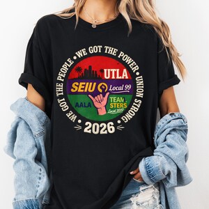 May include: Black t-shirt with a circular graphic featuring the text "WE GOT THE POWER" and "UNION STRONG." The graphic includes the text "UTLA," "SEIU Local 99," "TEAMSTERS Local 2010," and "2026." A hand gesture is also visible.