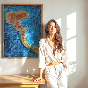 May include: An abstract painting in a wooden frame, featuring a map-like design in shades of orange and yellow against a blue background. The artwork hangs on a white wall, with a wooden table in front.