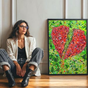 May include: A framed abstract painting of a broken heart. The artwork features a vibrant red heart split in two, set against a green and yellow background. The painting is in a black frame and leans against a white wall.