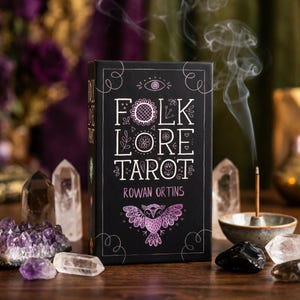 May include: A black tarot card box with the words "FOLK LORE TAROT" in white, and the author's name, Rowan Ortins. The box features a floral design and a purple owl illustration. Crystals and incense surround the box.
