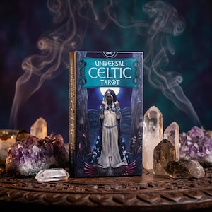 May include: A tarot card deck titled "Universal Celtic Tarot" with a teal and gold design. The box features an illustration of a robed figure. Clear and purple crystals surround the deck on a wooden surface, with wisps of smoke in the background.