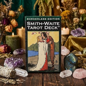 Puede incluir: Una caja de la baraja Smith-Waite Tarot Deck, edición sin bordes, con una ilustración de una pareja abrazándose. La caja está rodeada de velas, cristales, flores secas y salvia sobre una superficie de madera. El texto de la caja dice "Smith-Waite Tarot Deck".