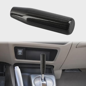 May include: A black carbon fiber shift knob, cylindrical in shape, is shown against a neutral background. The shift knob is a replacement part for a vehicle's gear shift. The image also shows the shift knob installed in a car's interior.