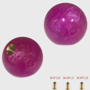 Peut inclure: Deux pommeaux de levier de vitesses sphériques magenta avec une finition nacrée. L'un des pommeaux a un insert fileté en laiton. Trois inserts en laiton de différentes tailles de filetage sont présentés en dessous. Accessoires automobiles.