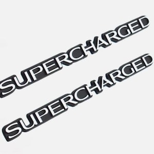 May include: Two black and white "SUPERCHARGED" emblems. The word "SUPERCHARGED" is in white, bold, capital letters on a black rectangular background. The emblems are angled on a white background.