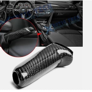 Puede incluir: Un pomo de palanca de cambios de fibra de carbono, con un acabado brillante, se muestra sobre un fondo interior de coche. El pomo es cilíndrico con un patrón texturizado. Un primer plano muestra el freno de mano de fibra de carbono.