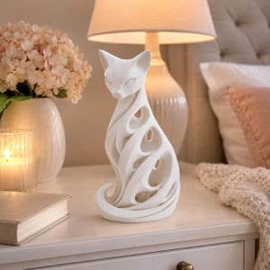 May include: A white cat figurine with an open, flowing design. The cat is sitting upright with its tail curled. The figurine is on a white surface, next to a lit candle, a book, and a vase of flowers. A lamp and a bed are in the background.