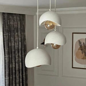 Op de afbeelding: Een witte, moderne hanglamp met drie koepelvormige kappen. Elke kap bevat een warmgekleurde gloeilamp. De lamp hangt aan een witte plafondbevestiging, wat een eigentijds verlichtingsontwerp creëert.