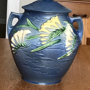 May include: A blue ceramic jar with a matching lid and two side handles. The exterior features a raised relief design of yellow and cream-colored flowers with green leaves against a textured, wavy background pattern that mimics water ripples.