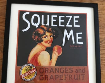 May include: A framed vintage-style advertisement print featuring a woman holding an orange. The text reads SQUEEZE ME BRAND, ORANGES and GRAPEFRUIT, and INTERNATIONAL FRUIT CORP. Balls of Juice, Lucerne Park, Florida. The design uses red, black, and white.