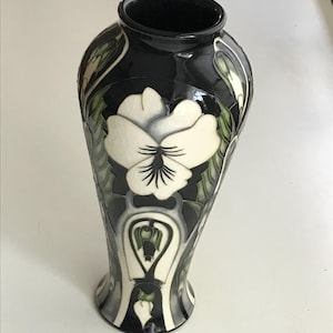 May include: A glossy black ceramic vase featuring a prominent white pansy flower design with green leaf accents. The surface has a visible crackle glaze finish, and the vase has a tapered neck with a wide opening and a flared base.