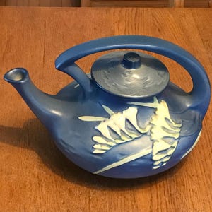 May include: A matte blue ceramic teapot featuring a raised, light-colored relief design of stylized floral elements. The teapot has a curved handle that connects to the lid area, a tapered spout, and a circular lid with a small central knob.