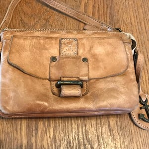May include: A tan leather crossbody bag featuring a front flap with a rectangular leather tab, dark metal hardware, and a matching leather strap. A circular leather tag with a dark metal anchor charm hangs from the side of the bag.