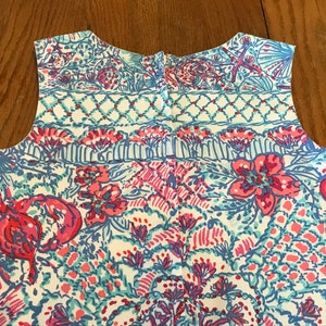 以下が含まれることがあります： 鮮やかな模様のノースリーブの衣服。 ピンク、青、ターコイズブルーの海のテーマのイラストが組み合わされており、花、貝殻、ロブスターなどが白い背景に描かれています。 生地は薄手に見えます。