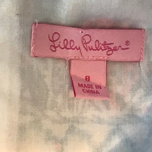 以下が含まれることがあります： ピンク色のラベル。筆記体のLilly Pulitzerのロゴが特徴。ロゴの下には、数字の8と「MADE IN CHINA」の文字が入った小さなタグがあります。白と青の模様の生地に配置されています。