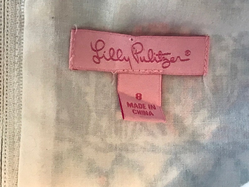 以下が含まれることがあります： ピンク色のラベル。筆記体のLilly Pulitzerのロゴが特徴。ロゴの下には、数字の8と「MADE IN CHINA」の文字が入った小さなタグがあります。白と青の模様の生地に配置されています。