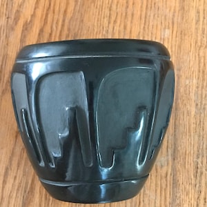 May include: A glossy black ceramic pot with a unique design. The pot features a series of carved, abstract shapes around its exterior. The pot has a rounded base and a slightly flared top, giving it a modern look. The pot is sitting on a wooden surface.