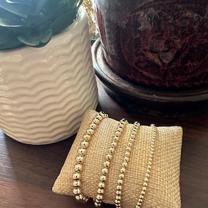 May include: Four gold beaded bracelets of varying sizes are displayed on a small, tan burlap pillow. The bracelets are made of shiny, round beads. The background includes a white ceramic pot with a plant and a dark brown pot.