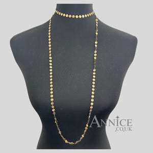 May include: A gold-coloured necklace and choker set displayed on a black mannequin. The necklaces feature a delicate design of small, round, flat discs. The long necklace hangs down the mannequin's front, while the choker sits at the neck.