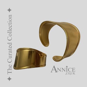 May include: Two gold-coloured metal cuff bracelets. One is a wide, curved design, and the other is a smaller, rectangular shape. The bracelets are displayed on a light grey background. Text on the left says "The Curated Collection" and the text "ANNICE .CO.UK" is on the bottom right.