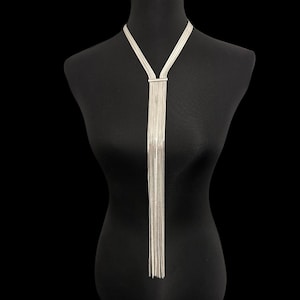 Op de afbeelding: Zilverkleurige ketting met een Y-vormig ontwerp. De ketting heeft meerdere strengen van delicate kettingen die naar beneden vallen vanaf een rechthoekige sluiting. De sieraden worden getoond op een zwarte mannequin tegen een zwarte achtergrond.