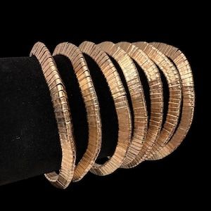 May include: Several gold-coloured, textured bracelets are displayed against a black backdrop. The bracelets feature a ribbed design and are arranged in a curved shape, demonstrating their flexibility. The solid black background accentuates the jewellery.