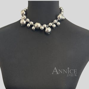 May include: A silver-toned choker necklace featuring a cluster of polished spherical beads of varying sizes suspended from a central chain. The necklace is displayed on a black mannequin bust with the text Annice.co.uk visible in the bottom right corner.