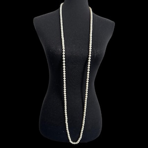 Puede incluir: Un collar largo de perlas blancas sobre un maniquí negro. Las perlas son pequeñas y están espaciadas uniformemente, creando un aspecto clásico y elegante. El collar cuelga por el torso del maniquí sobre un fondo negro.