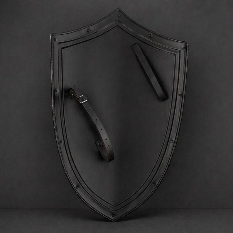 May include: A black shield with a pointed bottom and rounded top. The shield has a dark, matte finish and is edged with a raised border. Two black leather straps are attached to the back for holding. Rivets secure the border and straps.