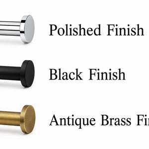 K&ouml;nnte beinhalten: Drei zylindrische Wandhaken mit flachen, runden Enden, die in einer vertikalen Reihe angeordnet sind. Der obere Haken hat ein gl&auml;nzendes, silbernes Polished Finish, der mittlere ein mattes Black Finish und der untere ein metallisches Antique Brass Finish.