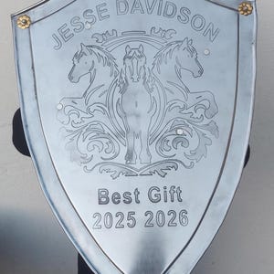 Puede incluir: Un escudo plateado con el nombre "Jesse Davidson" grabado en la parte superior. El escudo presenta un diseño de caballo y las palabras "Best Gift 2025 2026". El escudo tiene detalles florales dorados.