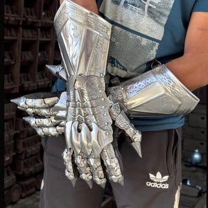 May include: A pair of silver-colored, articulated gauntlets. The metal gloves feature intricate engraved designs and pointed extensions at the knuckles and wrist. The gauntlets are designed to protect the hands and forearms.