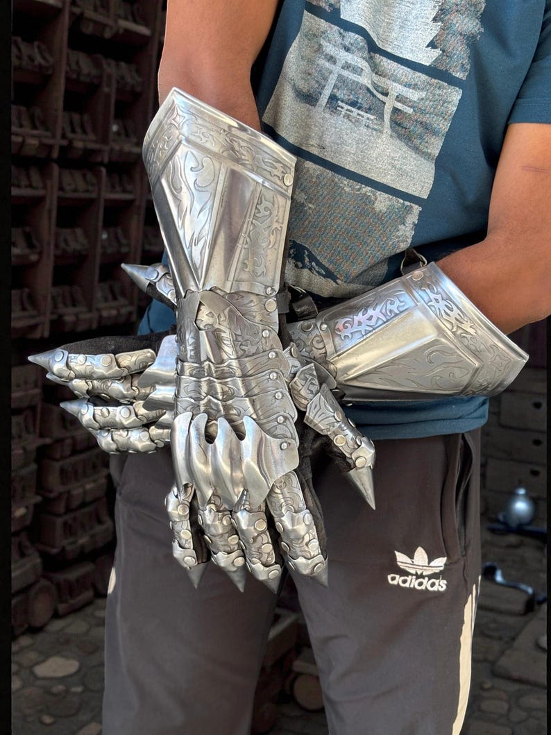 May include: A pair of silver-colored, articulated gauntlets. The metal gloves feature intricate engraved designs and pointed extensions at the knuckles and wrist. The gauntlets are designed to protect the hands and forearms.