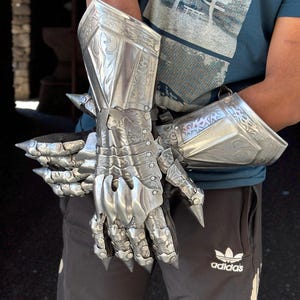 May include: Silver-colored, articulated gauntlets with intricate engraved designs. The armor features pointed extensions on the knuckles and forearms. The gauntlets are designed to cover the hands and forearms, offering a medieval aesthetic.