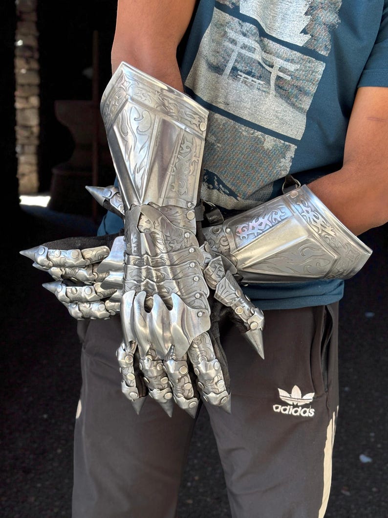 May include: Silver-colored, articulated gauntlets with intricate engraved designs. The armor features pointed extensions on the knuckles and forearms. The gauntlets are designed to cover the hands and forearms, offering a medieval aesthetic.