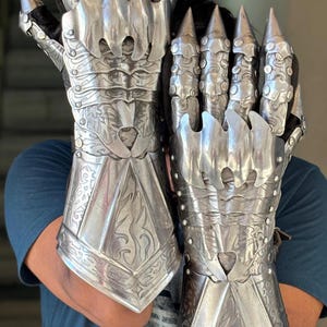 May include: A pair of silver-colored gauntlets with articulated fingers and sharp, conical tips. The metal gloves feature intricate engraved floral and geometric patterns, with heart-shaped cutouts. The gauntlets are designed to cover the entire hand and wrist.