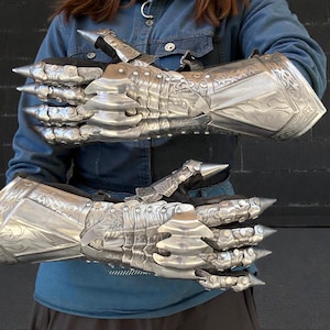 May include: A pair of silver-coloured, articulated gauntlets with sharp, pointed extensions at the knuckles and fingertips. The gauntlets feature engraved decorative patterns and are designed to cover the entire hand and wrist.