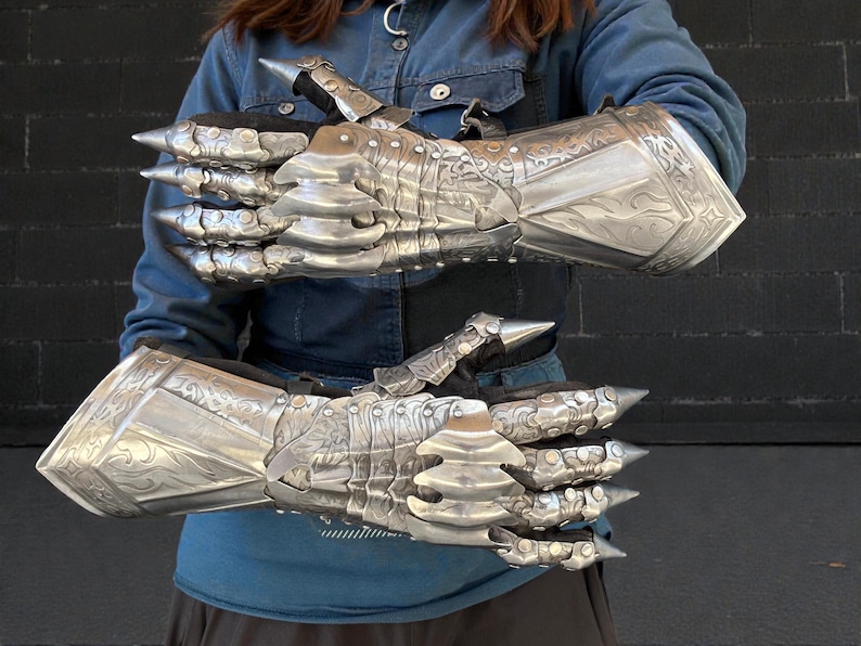 May include: A pair of silver-colored, articulated gauntlets with sharp, pointed extensions at the knuckles and fingertips. The gauntlets feature engraved decorative patterns and are designed to cover the entire hand and wrist.