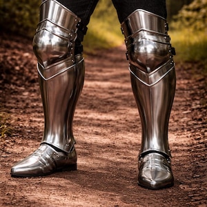 May include: A pair of polished steel leg armor pieces covering the lower legs and knees, featuring articulated knee caps and greaves that extend down to the feet. The metal plates are secured with black straps at the back of the knee joints.
