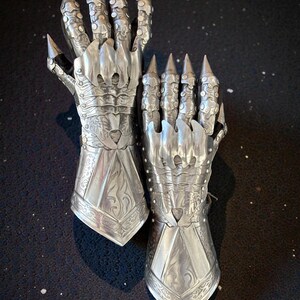 May include: A pair of silver-colored articulated gloves with pointed finger extensions. The gloves have intricate engraved designs and are made of metal. The gloves are designed to resemble claws.