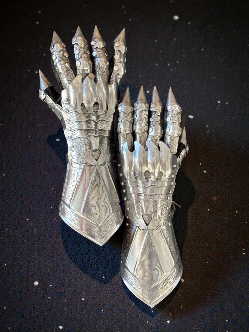 May include: A pair of silver-colored articulated gloves with pointed finger extensions. The gloves have intricate engraved designs and are made of metal. The gloves are designed to resemble claws.
