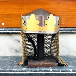 Könnte beinhalten: Ein mittelalterlicher Helm mit einem silberfarbenen, konischen Oberteil und einem goldfarbenen, kronenförmigen Band. Der Helm hat einen Kettenkragen und wird auf einem schwarzen Ständer mit rechteckiger Basis ausgestellt.