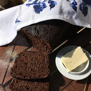 Könnte beinhalten: Ein Laib dunkelbraunes Brot, teilweise geschnitten, mit zwei Scheiben im Vordergrund. Ein kleiner weißer Teller enthält ein Stück Butter. Das Brot ist mit einem weißen Tuch bedeckt, das mit blauen Blumenmustern verziert ist.