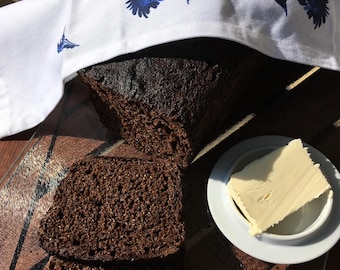 May include: A loaf of dark brown bread, partially sliced, with two slices in the foreground. A small white dish holds a pat of butter. The bread is covered with a white cloth decorated with blue floral patterns.