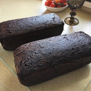 May include: Two dark brown loaves of bread on a glass board. A small bowl of cherry tomatoes and a decorative glass are in the background. The bread has a textured crust.