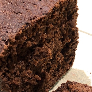 Könnte beinhalten: Nahaufnahme eines dunkelbraunen Brotlaibs mit rauer, strukturierter Oberfläche. Das Brot hat ein dichtes, feuchtes Aussehen mit sichtbaren Luftblasen. Eine Brotscheibe ist im Vordergrund zu sehen und zeigt die gleiche Textur und Farbe.