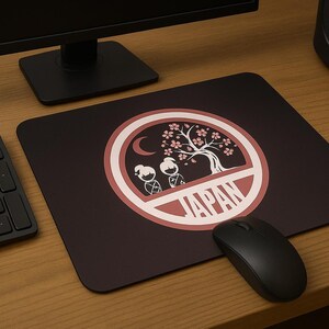 May include: A black computer mouse and keyboard sit on a wooden desk next to a black mousepad. The mousepad has a maroon background with a white and red circular design featuring a tree, figures, and the word "JAPAN."