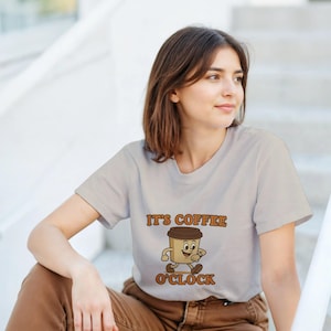Puede incluir: Una camiseta gris claro con el texto "IT'S COFFEE O'CLOCK" en letras marrones. La camiseta presenta un personaje de taza de café de dibujos animados con una cara sonriente y piernas caminando. La persona lleva pantalones marrones.