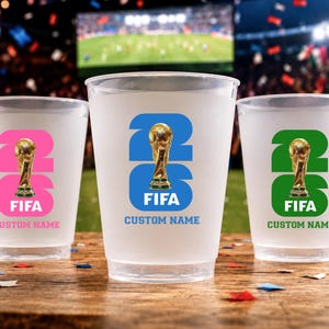 May include: Three frosted plastic cups with FIFA World Cup trophy designs. Each cup features a different colour scheme: pink, blue, and green. The text "26" and "FIFA" are displayed prominently, with space for a custom name.