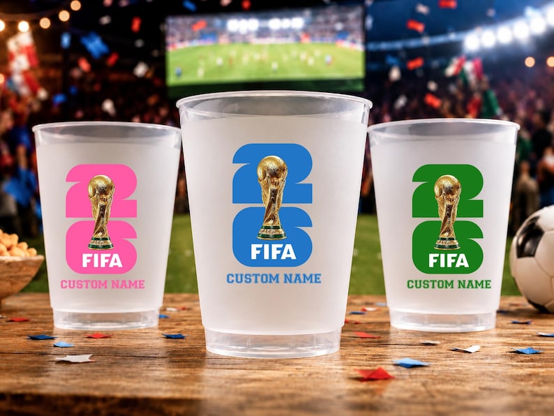 May include: Three frosted plastic cups with FIFA World Cup trophy designs. Each cup features a different colour scheme: pink, blue, and green. The text "26" and "FIFA" are displayed prominently, with space for a custom name.